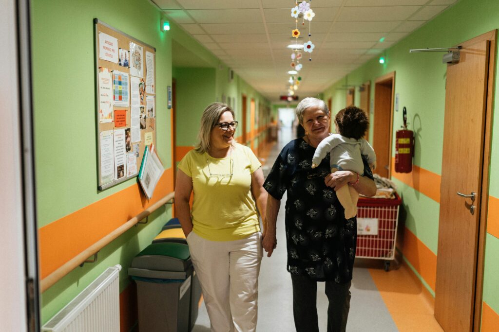 Two women walking in a nursing home corridor, one holding a child doll.